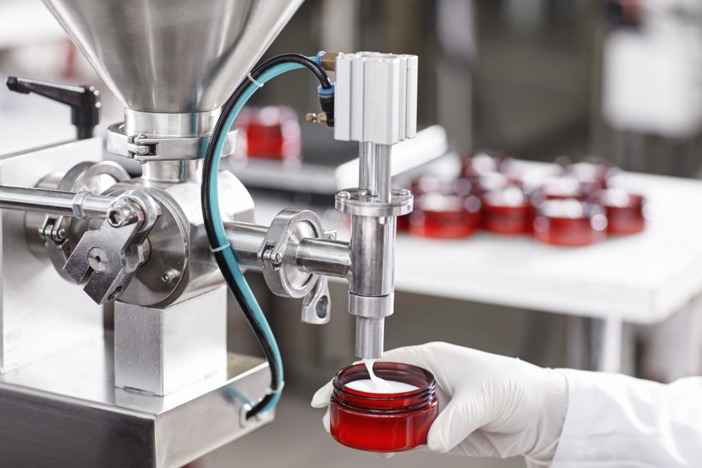 Hard mechanism of packing ready beauty cream into red container. A factory worker dressed in white lab gown and gloves holding red container under tap pouring ready cream. Production process of cream