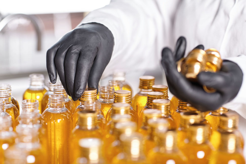 Selective focus of factory worker’s hands in black gloves closing bottles in front of him with screw caps at laboratory. Mass production and manufacturing. Medicine, cosmetics and pharmacy concept